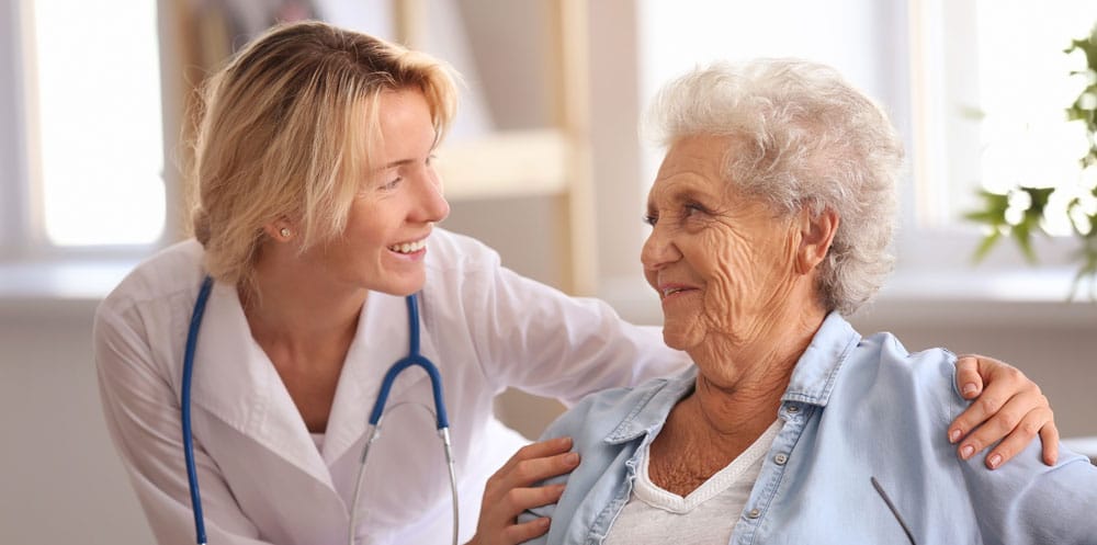 A senior woman is being cared for in a senior living community by a female doctor or nurse practitioner. The two women smile at each other while the doctor or nurse practitioner places her hands gently on the senior woman’s shoulders.