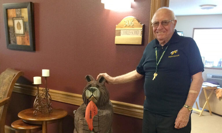 Senior male standing outside of his doorway next to a wooden bear carving.